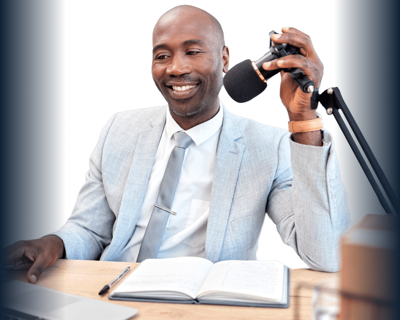 A smiling Black man in a professional gray suit adjusting a microphone at a desk with a laptop and open notebook.